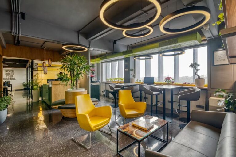 Un salon de bureau moderne avec des luminaires circulaires distinctifs au plafond, des fauteuils d’accent jaunes éclatants, un plafond industriel exposé, de grandes fenêtres, des espaces de travail à hauteur de bar et des plantes d’intérieur. L’espace combine des éléments commerciaux et confortables avec des touches de couleur.