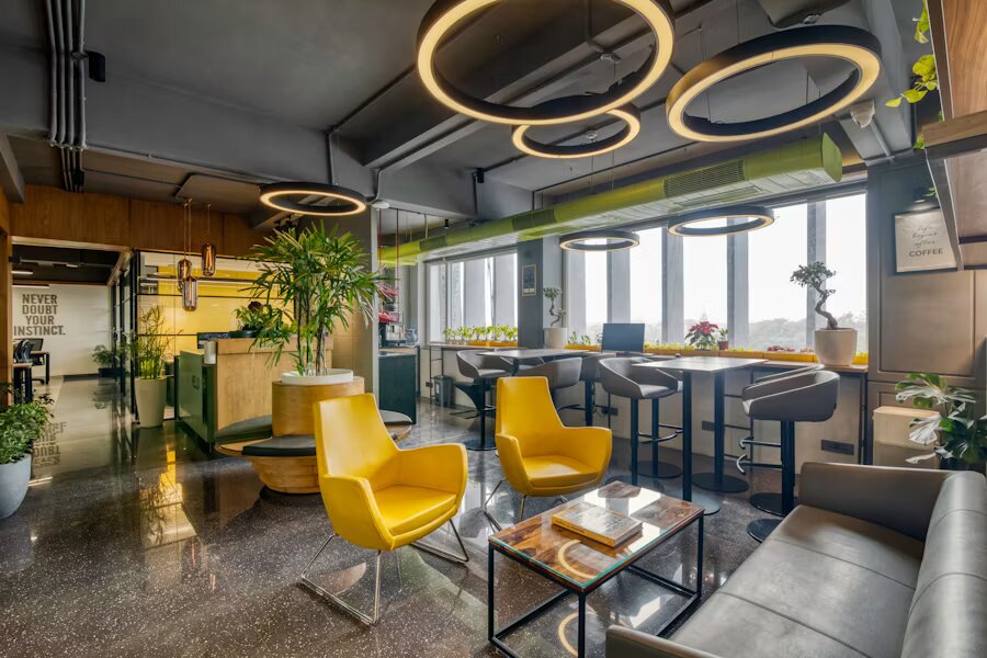 Un salon de bureau moderne avec des luminaires circulaires distinctifs au plafond, des fauteuils d’accent jaunes éclatants, un plafond industriel exposé, de grandes fenêtres, des espaces de travail à hauteur de bar et des plantes d’intérieur. L’espace combine des éléments commerciaux et confortables avec des touches de couleur.